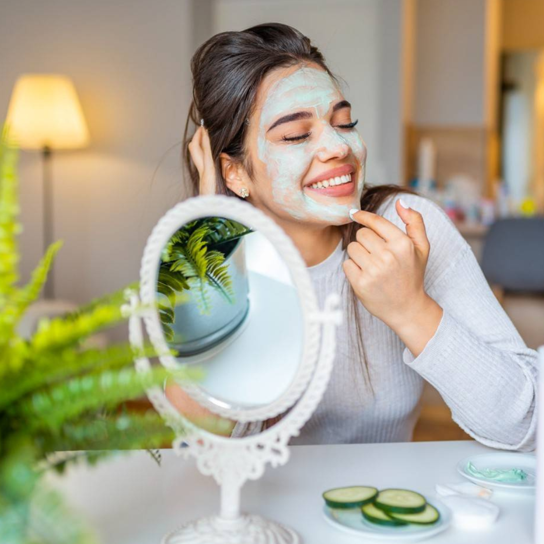 Woman applying a facial mask in front of a mirror with cucumbers on a table.