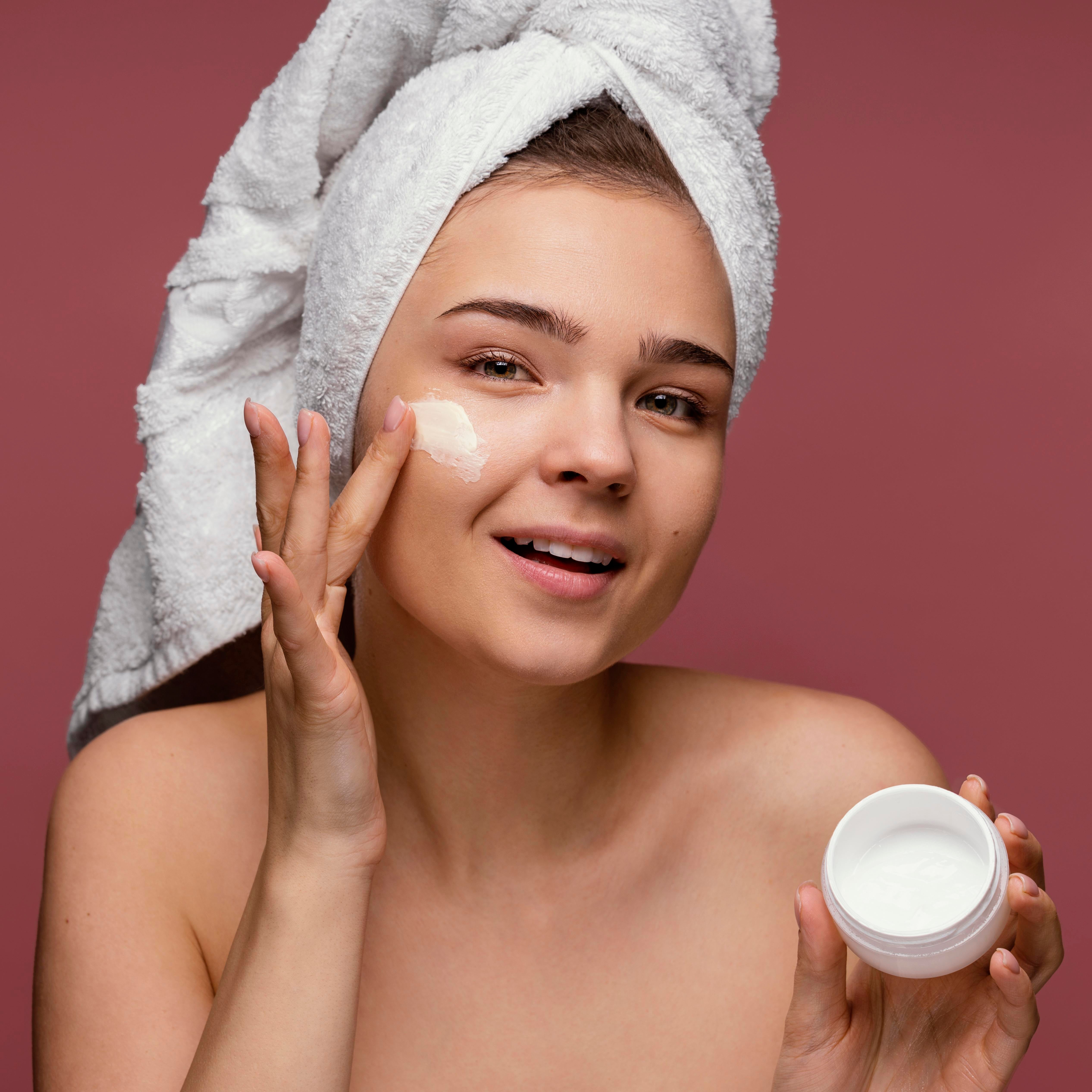 Woman applying cream to her face with a towel on her head against a maroon background